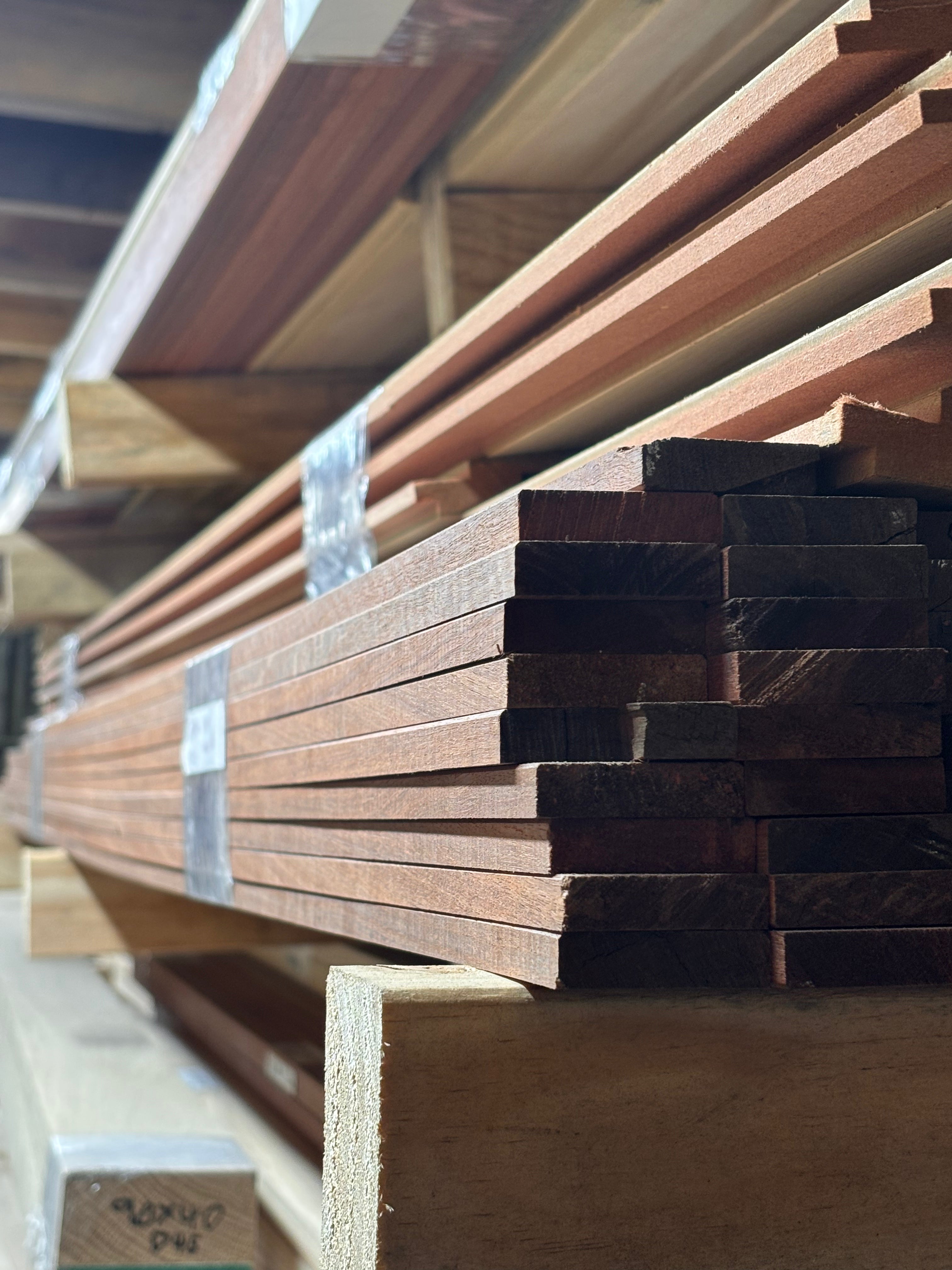 Stacks of raw wood planks and boards, including reddish-brown and dark brown timber, secured with clear plastic bands on wooden shelving.