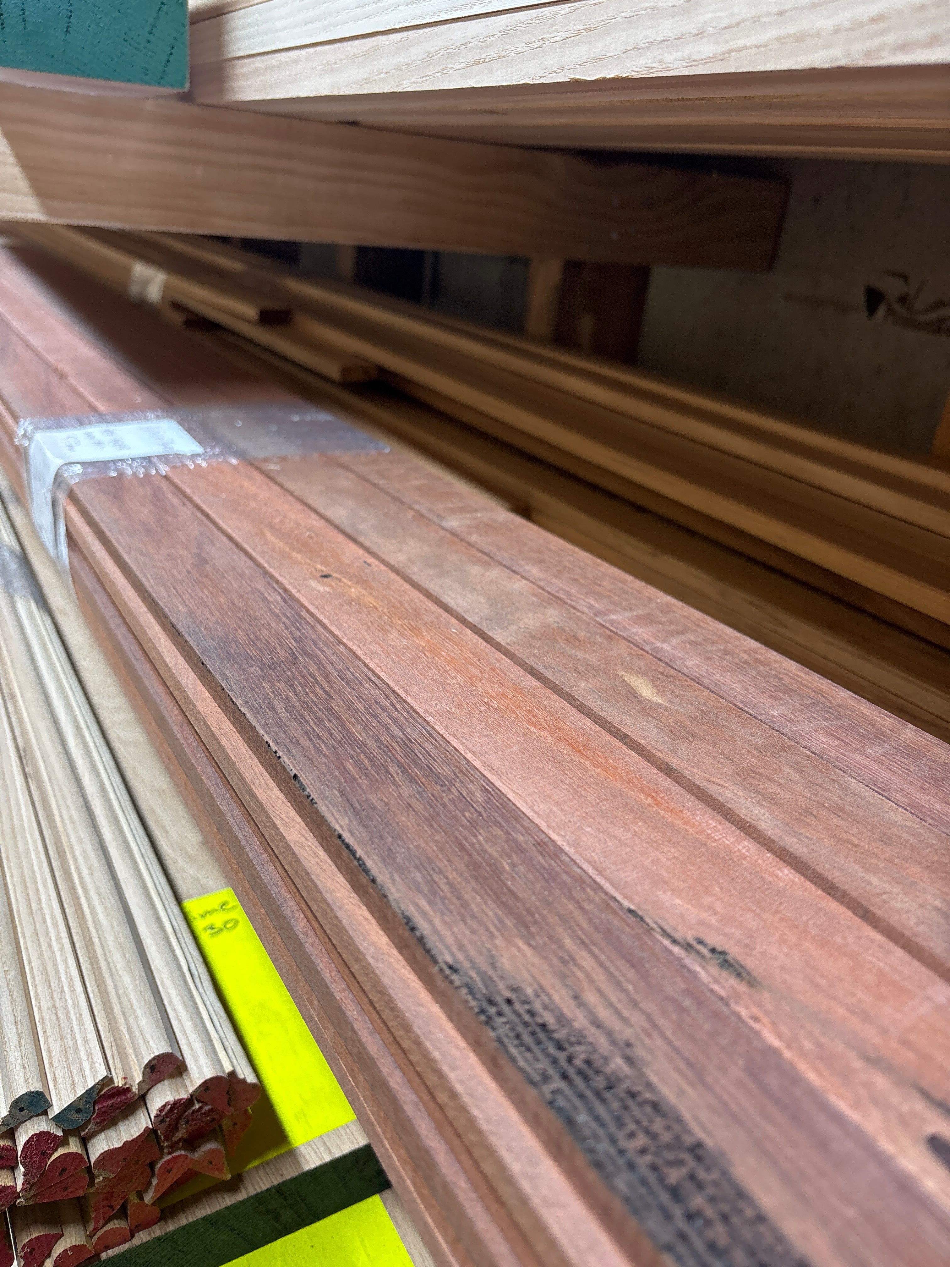 Close-up of a wooden board or trim piece with visible wood grain, photographed in a workshop or storage area with other lumber and a yellow measuring tape visible in the background.