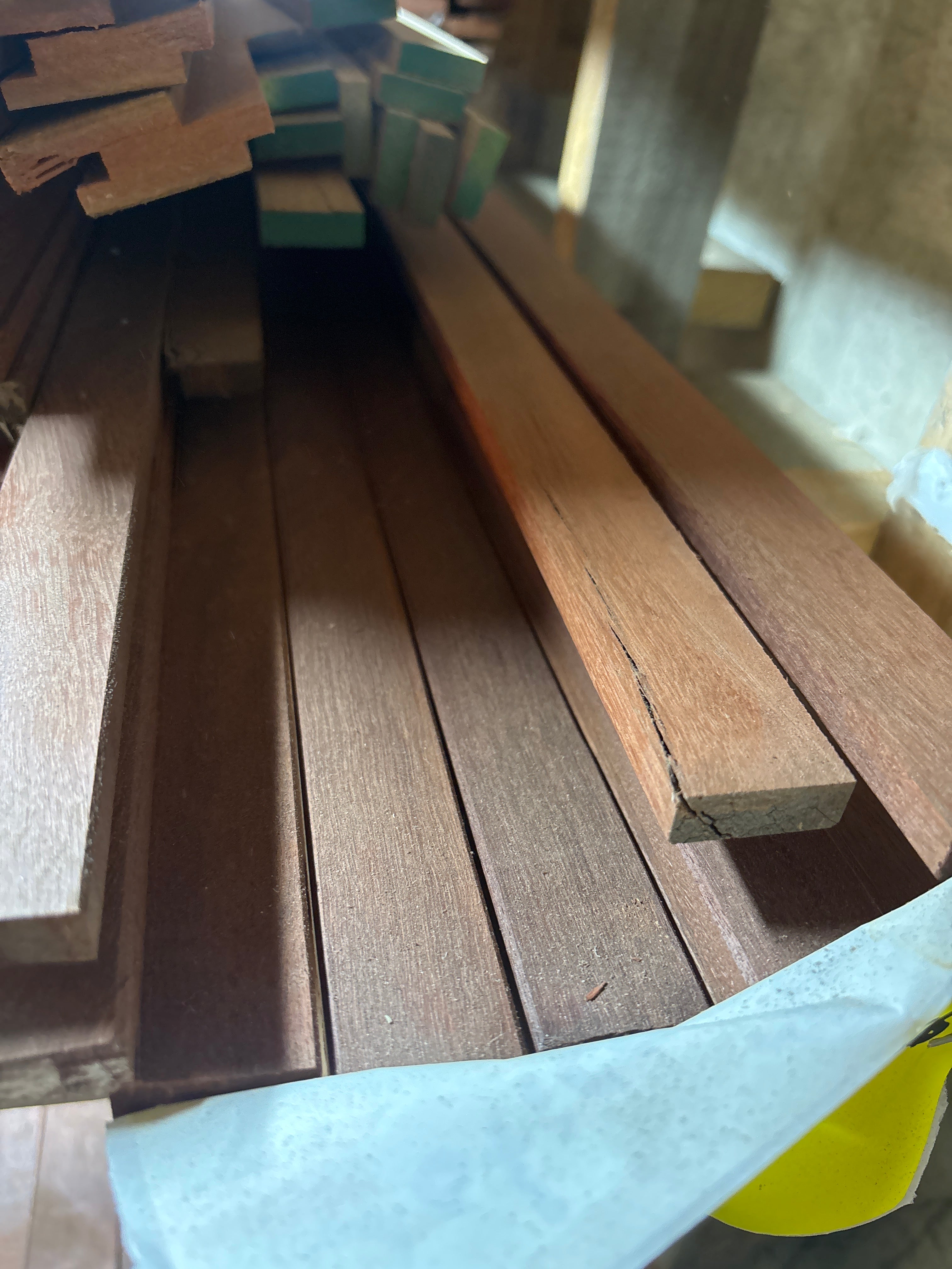 Stacks of dark brown wood planks with a visible grain and a small edge notch, partially covered by a light gray sheet of paper.